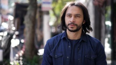 Pensive young casual man walking in street outdoors. Thoughtful person in 30s walks in urban environment. Male with long hair