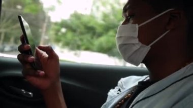 Black man in backseat car looking at cellphone screen wearing face mask prevention against pandemic