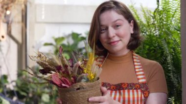 Portrait of a young employee showing bouquet arrangement at flower shop. Woman wearing apron holding flower in hand looking at camera