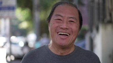 Happy Asian American senior man smiling at camera while walking outdoors. Excited middle aged Japanese person walks in urban street sidewalk