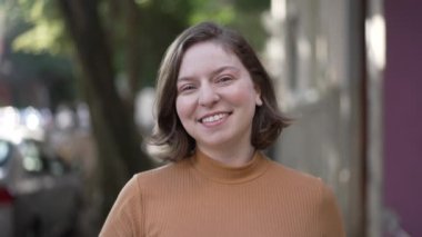 Portrait of a joyful woman walking toward camera. Tracking motion shot of person coming forward smiling. Happy millennial 20s girl in city sidewalk