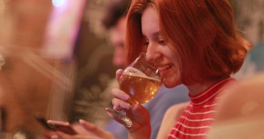 Samimi kızıl saçlı kadın cep telefonu ekranına bakarken bira içiyor.