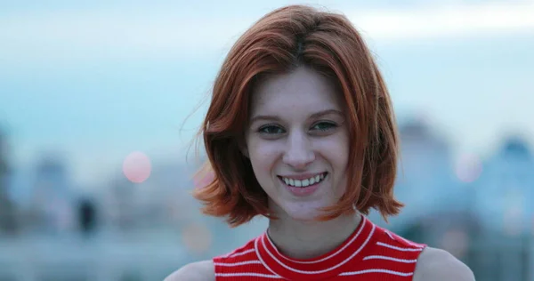 Portrait of young redhead millennial woman smiling to camera in city in background