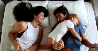 Millennial couple in bed with infant toddler boy wanting attention. Man and woman lying down looking at cellphone, top view