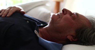 Older man lies in bed to rest, senior looks at camera while laying down