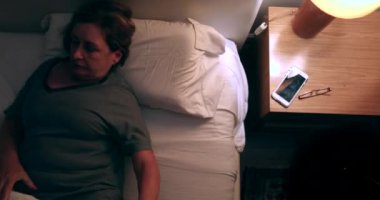 Older single woman lies down in bed yawning and picking up cellphone device. Putting reading glasses