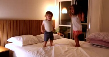 Happy children bounding up and down in bed at night. Little girl and toddler boy jumping in bed