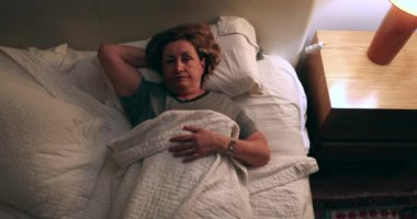 Pensive older woman lying down in bed, turns off nightstand light off and goes to sleep