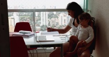 Mother multi-tasking at home, infant toddler boy watching cartoon on laptop while mom studying