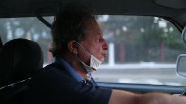Older man driving car wearing covid-19 face mask. Senior removing surgical mask while on road