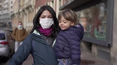 Mother wearing covid surgical face mask walking in street with toddler baby