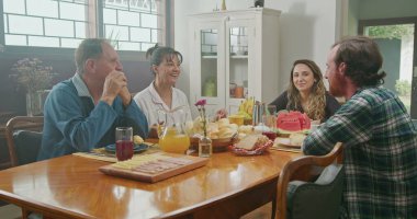 Orta yaşlı ebeveynler, küçük kızı ve büyük oğluyla kahvaltı yapıyor. Anne kocasına peynir ekmeği servis eder. Oğlu ekmek yiyor, mutlu mesut sohbet ediyorlar..