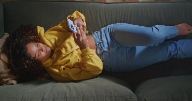 African American woman lying on sofa holding smartphone seen from above angle. A black girl taking selfie with phone