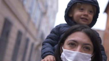 Mother wearing covid face mask walking outside with baby toddler in shoulders