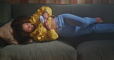 African American woman lying on sofa holding smartphone seen from above angle. A black girl taking selfie with phone