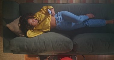 A black girl lying on a sofa using a smartphone. One African American young woman on a comfortable sofa holding cellphone. Relaxed happy woman lying on sofa and surfing internet. From above angle