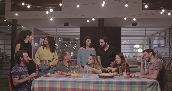 Group of happy people are sitting together in the backyard of the house at night laughing and smiling. Everyone is gathered around the table chatting happily.