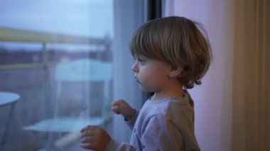 Bored little toddler child wanting to outside stuck at home. Baby standing by window looking out