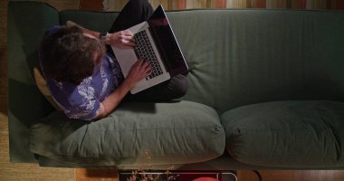 Oturma odasındaki kanepede oturan adam. Bilgisayarda internette gezinen kişinin yukarıdaki açısından