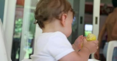 Parent feeding adorable baby infant on highchair. Cute toddler holding and eating corn