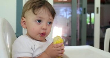 Parent feeding adorable baby infant on highchair. Cute toddler holding and eating corn