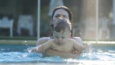 Anne yüzme havuzunda neşeli bir bebek tutuyor. Yavaş çekimde su sıçratan bebek 120 fps