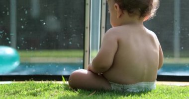 Dışarıda çimenlerin üzerinde oturan, arka planda havuzu olan güzel bir bebek.