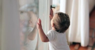 Baby standing by window wanting to leave. Cute infant toddler leaning on glass