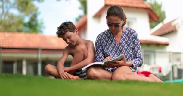 裏庭の外で子供の男の子と母親の読書 屋外で勉強する宿題を持つ少年を助ける親 — ストック動画