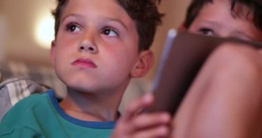 Çocuk elinde tablet tutarken TV ekranına bakıyor