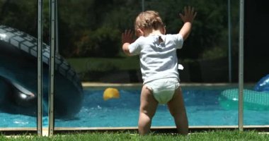 Yüzme havuzu çitlerine yaslanmış şirin bir bebek. Kardeşlerin suyun içinde oynamasını izliyor.