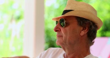 Older man wearing sunglasses and hat relaxing outside looking at the horizon resting