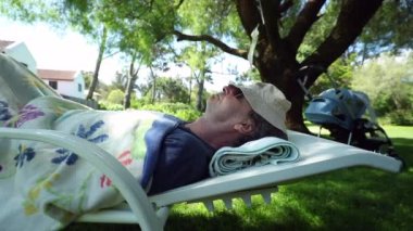 Older man resting outside under tree, candid senior retired person scratching ear and stretching body