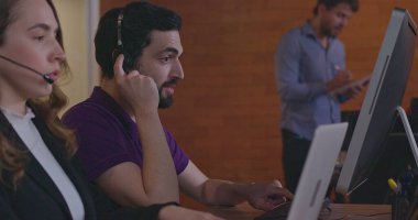 Tele pazarlamacılar dizüstü bilgisayarların önünde kulaklık takan müşterilerle iletişim kuruyor. Ofis içinde çalışan kadın ve erkek çalışanlar çağrı merkezindeki müşterilerle konuşuyor.