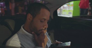 Person looking at cellphone riding taxi in backseat of car. African American person staring at smartphone screen after work in city