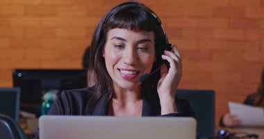 Ofis çağrı merkezinde bilgisayar ekranının önünde kulaklıkla konuşan kendinden emin genç bir kadın. Müşteri hizmetlerinde çalışan bayan çalışan müşteriyle çevrimiçi konuşuyor. Güney Amerikalı çalışanlar gece çalışma alanında