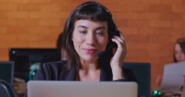 Ofis çağrı merkezinde bilgisayar ekranının önünde kulaklıkla konuşan kendinden emin genç bir kadın. Müşteri hizmetlerinde çalışan bayan çalışan müşteriyle çevrimiçi konuşuyor. Güney Amerikalı çalışanlar gece çalışma alanında