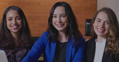 Üç iş kadını kameraya bakıp gülümsüyor. İş yerindeki dizüstü bilgisayarın önünde oturan bir grup bayan meslektaşım. Çalışanların portresi