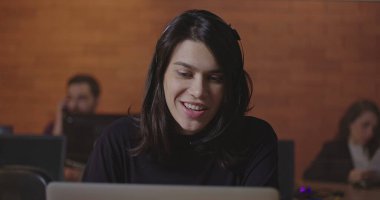 Bilgisayarın önünde konuşan kulaklık takan kadın bir ofis çalışanı. Genç bir kadın, iş yerindeki bilgisayar ekranının önünde gece vakti iş arkadaşıyla video üzerinden konuşuyor.