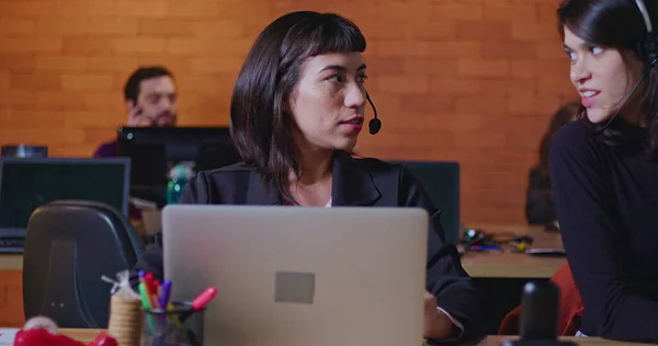 Female employee speaking with colleague at call center giving feedback. Two women wearing headset in communication in front of computer screens. People at workspace working at night