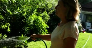 Woman in 60s watering plants with water hose outdoors with sunlight lens-flare