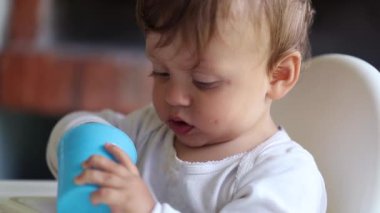 Sevimli erkek bebek sandalyeye oturmuş plastik mavi bardakla oynuyor.