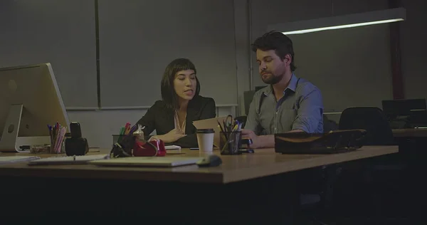 Two people working over time at office at night. Male and female employee sitting at workplace in the late evening revising documents and paperwork