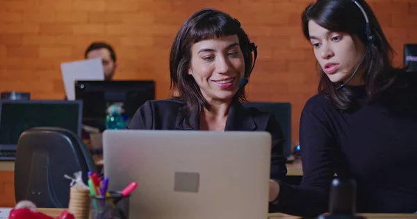 Female employee speaking with colleague at call center giving feedback. Two women wearing headset in communication in front of computer screens. People at workspace working at night