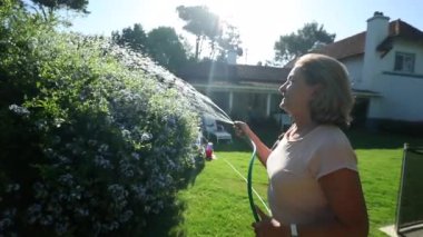 Senior woman watering plants with water hose. Older lady in 70s gardening and taking care of home backyard