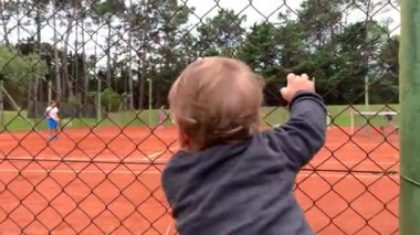 Tenis kortunun çitlerine tutunup oyun oynayan kardeşlere bakan sevimli bir bebek.