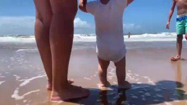 Bebek sahilde ilk kez yürüyor, anne kıyıda küçük bebekle el ele tutuşuyor ve yardım ediyor.