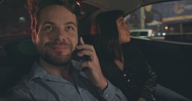 People riding taxi cab in car backseat at night. Young man and woman passengers in the evening after work talking on phone