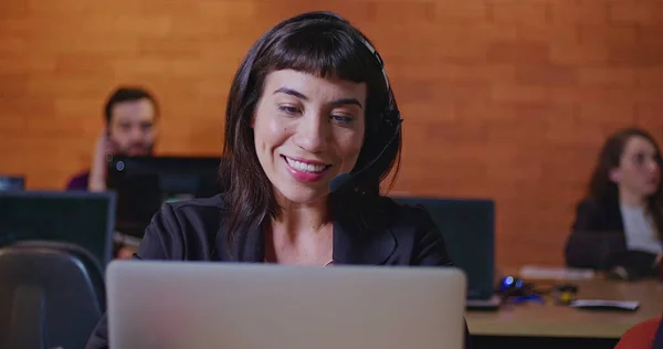 South American female employee speaking with headset in front of laptop. A hispanic latin young woman in communication via video conference speaking with client or customer inside corporate office workspace