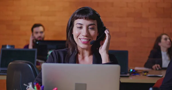 South American female employee speaking with headset in front of laptop. A hispanic latin young woman in communication via video conference speaking with client or customer inside corporate office workspace